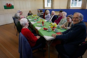 Enjoying the Bara Brith and Welsh Cakes at the St David's Day Guilsfield Gets Together Coffee Morning with daffodils on the table and a Welsh flag on the wall
